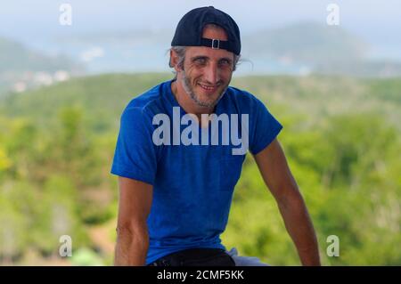 Felice giovane uomo barbuto seduto sulla pietra ringhiere in city park indossa nel bianco blu t-shirt e tappo nero Foto Stock