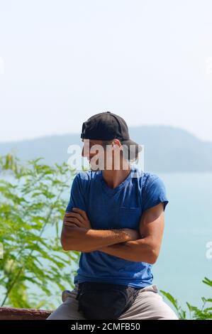 Felice giovane uomo barbuto seduto sulla pietra ringhiere in city park indossa nel bianco blu t-shirt e tappo nero Foto Stock