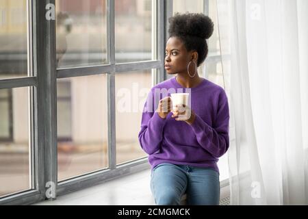 La donna biraciale millenaria afro-americana con orecchini di gioielli alla moda indossano il maglione viola pensando, guardando attraverso la finestra, tenendo il whit Foto Stock