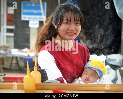 Bella giovane donna vietnamita tiene il suo bambino addormentato tra le braccia e guarda in uno specchio sporco e offuscato barbiere-negozio. Foto Stock