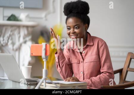 Donna millennial afroamericana con studio remoto afro-acconciatura, lavorando online su laptop, chattando con gli amici tramite videochiamata su smartphone Foto Stock