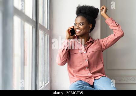Felice donna millennial afroamericana con acconciatura afro indossare camicia rosa, seduta sul davanzale, sorridente, prendendo il telefono, guardando la finestra. Nero Foto Stock