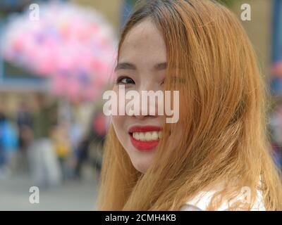 Giovane donna vietnamita con capelli tinti e rossetto rosso guarda sopra la spalla e sorride per la fotocamera. Foto Stock