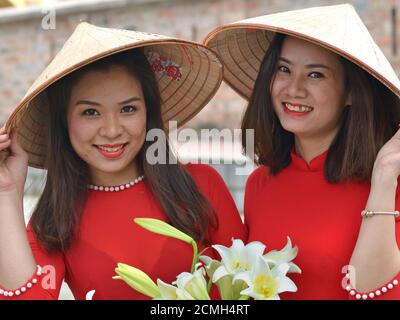 Due giovani donne vietnamite indossano cappelli conici asiatici e abiti rossi vietnamiti, tengono con le mani brim dei loro cappelli e sorridi per la macchina fotografica. Foto Stock