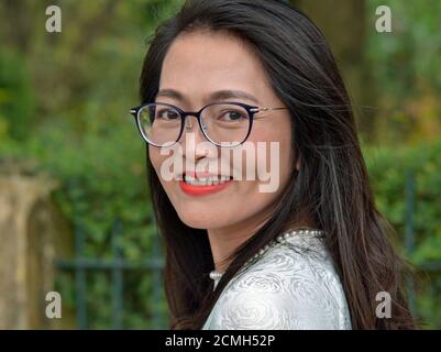 Giovane donna asiatica d'affari con i capelli lunghi e gli occhiali moderni guarda sopra la sua spalla e sorride per la macchina fotografica. Foto Stock