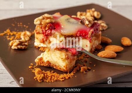 Una fetta di cheesecake di frutta servita su un piatto di porcellana marrone con noci e mandorle. La torta ha lampone e kiwi che è rifinito con Foto Stock