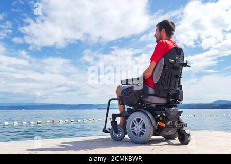 Uomo con distrofia muscolare su sedia a rotelle elettrica all'aperto guardando il mare. Foto Stock