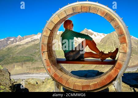 Donna escursionista che si rilassa all'interno della panca di legno dopo il trekking nella catena montuosa delle Alpi Bernesi svizzere, Canton Vallese, Europa. Donna che guarda il ghiacciaio di Aletsch Foto Stock