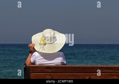 Donna in cappello da sole seduta su una panchina di legno su sfondo blu del mare. Vacanza in spiaggia, godendo del concetto di vita, relax e svago Foto Stock