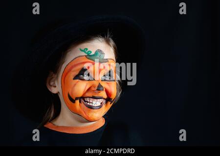 Ragazza in un cappello nero e trucco di zucca per Halloween guardare lateralmente su uno sfondo nero, simulate in su Foto Stock