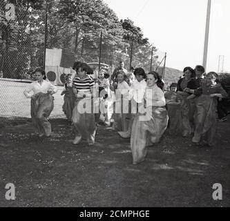 Anni '60, storica, estiva e all'aperto su una zona di erba, ragazze di diverse età più un paio di mamme, partecipando a una gara di sacco a una scuola fete, Bowhill, Scozia. Con le gambe all'interno di un sacco, i concorrenti saltano in avanti o, a volte, come si vede qui, a seconda delle dimensioni dei sacchi, corrono in loro su una distanza predeterminata. Un'attività informale ma tradizionale e divertente che si svolge nelle giornate degli sport scolastici e della carità. Foto Stock