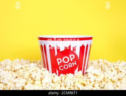 Primo piano di una benna di popcorn a strisce rosse e bianche circondata da popcorn appena schioccati, sfondo giallo brillante con spazio per la copia. Foto Stock