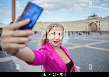 La donna bionda caucasica di mezza età in tuta rosa sta prendendo selfie dal telefono cellulare nel centro storico di San Pietroburgo, Russia. Foto Stock