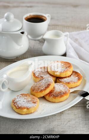 La prima colazione. Frittelle di formaggio con panna acida spolverati con zucchero a velo su una piastra bianca. Foto Stock