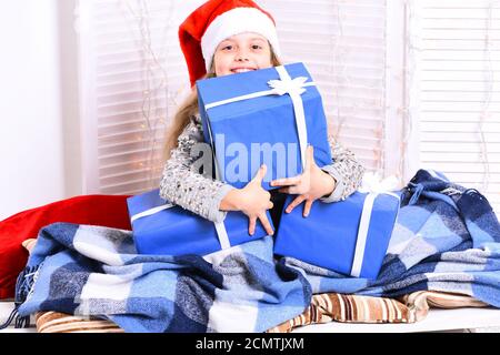 Ragazza con faccia allegra si siede su sfondo chiaro finestra. Natale e concetto di infanzia. Santas helper in cappello rosso coperto di coperta blu contiene scatole regalo blu. Il bambino si prepara alle vacanze invernali Foto Stock