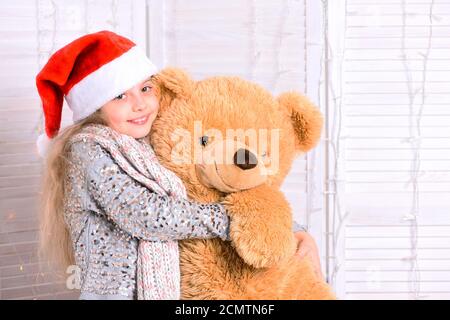 Miss Babbo Natale in cappello rosso abbracci enorme orso giocattolo. Ragazza con faccia sorridente posa su sfondo chiaro finestra. Vacanze invernali e concetto di infanzia. Il bambino ottiene il regalo di Natale. Foto Stock