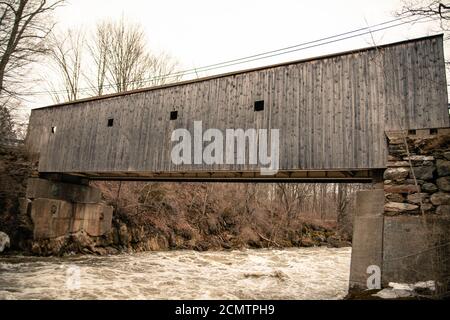 Il ponte rustico coperto è una struttura in legno-capriate sopra fiume rapido durante un'inondazione in una cupa giornata grigia nei vecchi colori del seppia Foto Stock