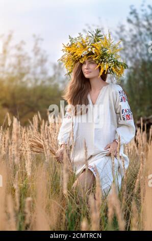 Giovane bella donna in una corona di fiori selvatici e antica camicia ricamata in un prato di erba secca gialla. Cultura Ucraina, stile etno Foto Stock