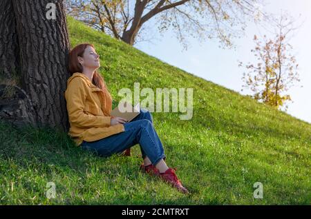 Bella e felice giovane donna gode di una piacevole seduta serale sull'erba da un albero in una molla o. parco autunnale Foto Stock