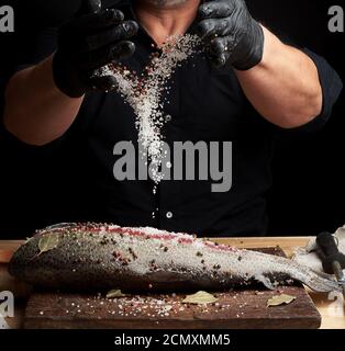 chef in una camicia nera e guanti in lattice nero prepara filetto di salmone su un tagliere di legno Foto Stock