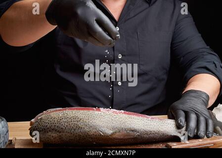 chef in una camicia nera e guanti in lattice nero prepara filetto di salmone su un tagliere di legno Foto Stock