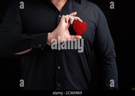 l'uomo adulto si erge su uno sfondo scuro con un colore nero camicia e tenendo un cuore rosso intagliato vicino al suo Foto Stock