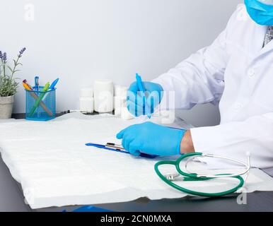 medico in un cappotto medico bianco si siede a un tavolo e scrive con una penna su un foglio bianco di carta Foto Stock