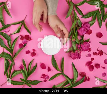 due mani di una giovane ragazza con pelle liscia e. un vasetto con crema spessa e peonie di borgogna Foto Stock