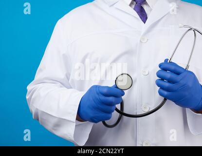 medico maschile adulto in un cappotto medico bianco stand e. contiene uno stetoscopio nero su sfondo blu Foto Stock