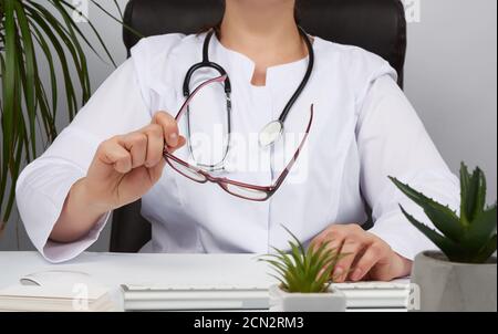 donna medico in una uniforme bianca, maschera medica monouso si siede in una sedia al tavolo e funziona Foto Stock
