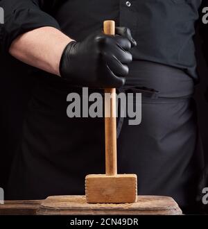 cuoco in uniforme nera tiene un martello di legno per battere carne su un tagliere vintage Foto Stock