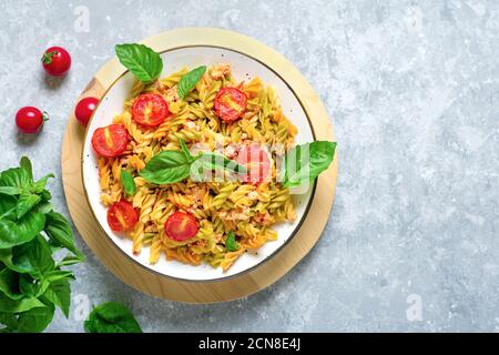 Fusilli - classica pasta italiana di grano duro con carne di pollo, pomodori ciliegia, basilico in salsa di pomodoro in ciotola bianca su tavola grigia in cemento Foto Stock