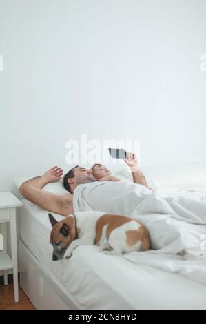 Papà e figlia che guardano lo smartphone a letto con il cane Foto Stock