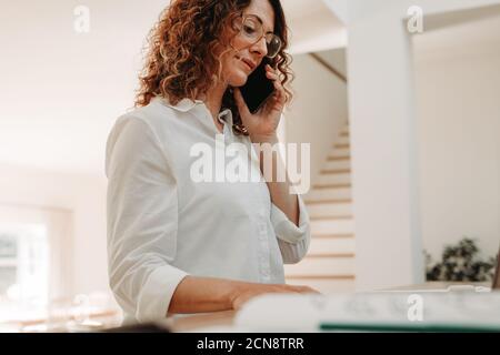 Donna seduta a casa che effettua una telefonata mentre lavora su un laptop. donna d'affari con capelli ricci che indossa occhiali che lavora da casa. Foto Stock