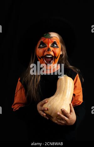 un bambino in un cappello nero e trucco di zucca urla su uno sfondo nero tiene una zucca nelle mani e ride Foto Stock