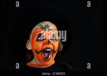 un bambino in un cappello nero e trucco di zucca urla su uno sfondo nero, isolato Foto Stock