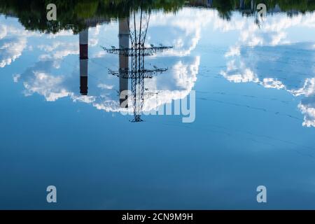 Riflesso di una linea elettrica, due alte tubature e nubi di cumulo scuro in acqua in una giornata estiva. Foto Stock
