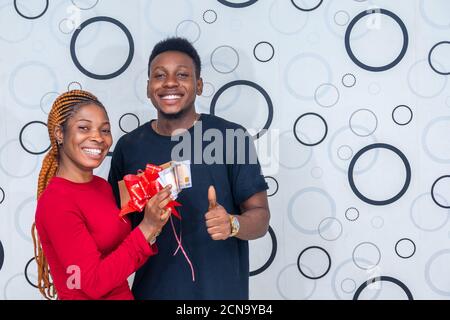 donna eccitata che sfalcia un avvolto con un nastro contenente regalo in contanti e un ragazzo che sta accanto al suo sorriso e dà un pollice in su Foto Stock