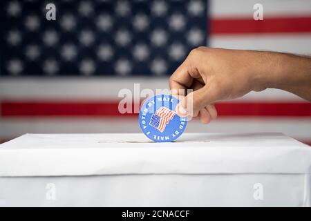 Primo piano di mani che cadono ho votato i miei adesivi Mail All'interno della casella di voto con la bandiera USA come sfondo - Concetto di voto per posta o posta in votazione all'indirizzo Foto Stock