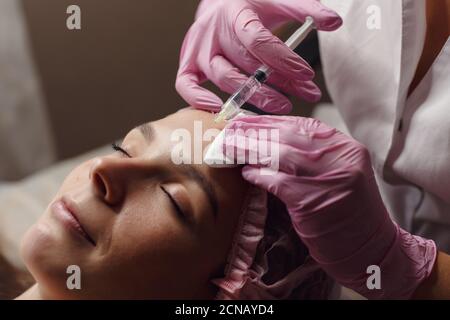 Donna sulla procedura di ringiovanimento in iniezione di riempimento di clinica di bellezza. Lei sta sdraiato tranquillamente in clinica. L'esperto estetista in guanti rosa sta riempiendo la donna Foto Stock