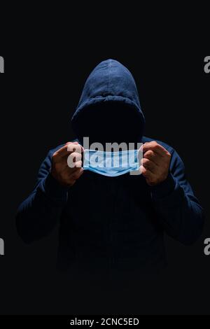Uomo senza volto in blu scuro felpa con cappuccio che mette su maschera chirurgica blu, faccia nascosta in ombra, buio intorno a lui Foto Stock