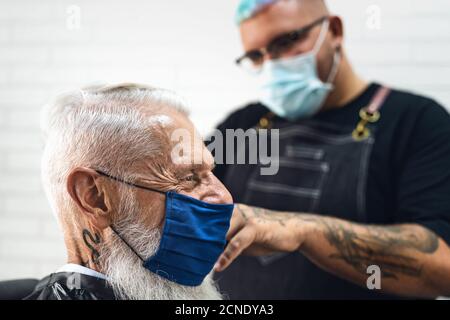 Parrucchiere maschile taglio capelli per hipster senior client mentre si indossa Maschera chirurgica facciale - giovane hairstylist che lavora in barbershop durante corona virus Foto Stock