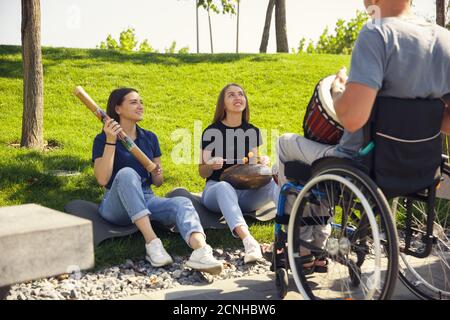 Ispirazione. Felice uomo caucasico portatore di handicap su una sedia a rotelle trascorrere del tempo con gli amici giocando musica strumentale dal vivo all'aperto. Concetto di vita sociale, amicizia, possibilità, inclusione, diversità. Foto Stock