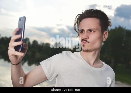 frustrato giovane bel ragazzo fa fallimentare selfie sul suo smartphone in natura, il ragazzo grimaces nella fotocamera.l'uomo re Foto Stock