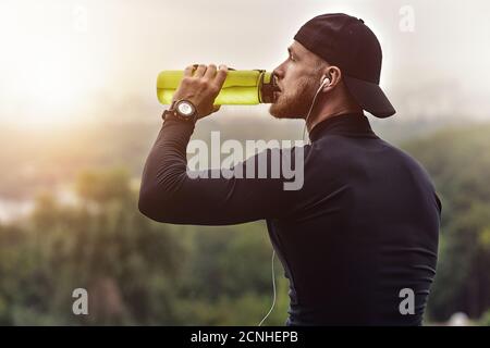 Muscolare atleta barbuto bere un acqua dopo una buona sessione di allenamento sul parco della città. Foto Stock