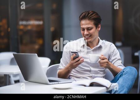 Giovane uomo sicuro con un abbigliamento casual elegante che tiene una tazza e parla con uno smartphone. Foto Stock