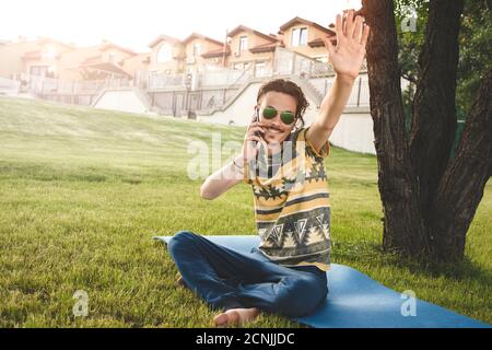 giovane ed elegante uomo sorridente con occhiali da sole è seduto sull'erba, riposandosi e parlando al telefono Foto Stock
