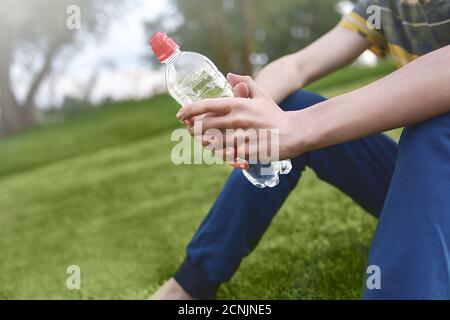 Giovane uomo caucasico corridore rilassante tenendo bottiglia di acqua potabile e. seduto sull'erba nel parco all'aperto dopo lo sport a. presto Foto Stock