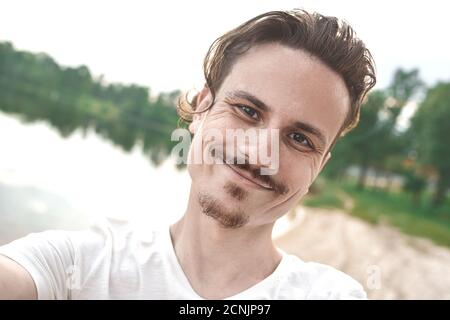 Bel ragazzo caucasico sorridente prende un selfie alla spiaggia - persone, stile di vita. Guardando nella macchina fotografica Foto Stock
