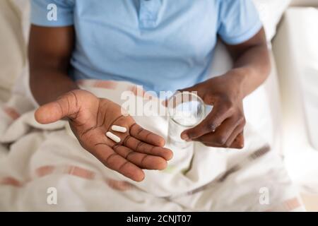 L'uomo anziano afroamericano seduto su un letto in una camera da letto, tenendo due pillole mediche e un bicchiere d'acqua nelle sue mani, l'allontanamento sociale e l'iso di auto Foto Stock